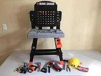 Front view of the child's Black & Decker workbench, showing pegboard, work surface, various toy tools, safety helmet, and goggles arranged in front.