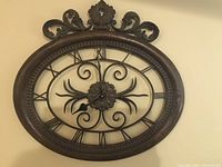 Front view of an oval metal wall clock with ornate top decoration, roman numerals and central floral scrollwork.
