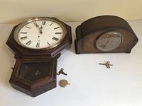 Photo showing two wooden clocks side by side with winding keys and pendulum on white surface.