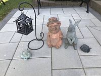 Photo showing front view of pig statue, rabbit, two frog figurines and lantern stand with hanging lantern on paved ground.