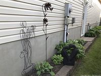 Photo showing the 4 metal garden ornaments placed on ground beside house siding, including wire man/woman figure, floral metal stake, black abstract heart sculpture, and another floral metal stake.