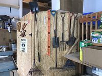 Photo shows nine assorted garden tools hanging on a particle board wall inside a workshop or shed. Tools include shovels, rakes, pitchforks, and a level.