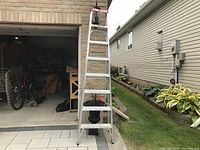Full front view of the open 8 ft aluminum step ladder with visible steps and red top cap, standing on tiled area in front of garage and garden.