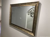 Front angled view of large rectangular mirror mounted on wall showing the textured gold-toned frame and beveled glass reflectance.