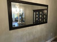 Rectangular mirror mounted on a wall reflecting a chandelier, furniture, and a display cabinet in the background. The black frame has a subtle inner bevel detail.