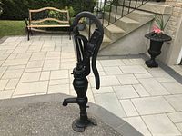 Full side view of old cast iron hand water pump showing lever handle, spout, and mounting base on pavement and driveway.