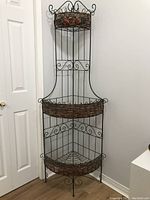 Full view of the tall, three tier metal corner plant stand with woven basket shelves and decorative metal roses.