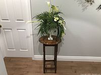 Full view of solid wood tall plant stand with round decorative top and silk plant arrangement with white flowers and green leaves.