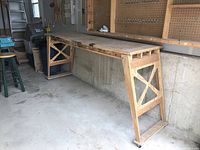 Side view of long homemade wooden work table on wheels showing wooden legs with wheel on one side and solid top surface with visible wear.