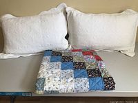 Two white quilted pillows with folded patchwork quilt in front on a table.