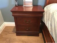 Front view of one of the wooden end tables showing two drawers with brass handles, dark cherry finish, next to a bed.