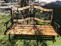 Front view on grass under tree, shows black wrought iron frame with wooden slat seat and floral backrest.