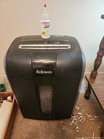 Front view of large black Fellowes paper shredder with visible shredded paper inside the transparent window and shredder lubricant bottle on top.
