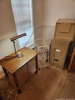 View of small wooden table on wheels with a metal desk lamp on top, white wire cart adjacent to filing cabinets.