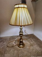 Full view of the brass lamp with beige pleated shade showing entire lamp and cord on floor.