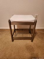 Front view of small vanity stool showing white cushioned seat, brushed chrome frame, and lower slatted storage shelf.