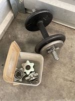 Adjustable dumbbell with black and silver metal plates and handle alongside a translucent container holding star-shaped metal weight collars.