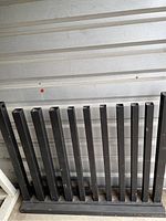 Photo showing the disassembled black metal dumbbell rack parts with vertical slots for dumbbells against a wall.