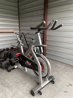 Front-side view of spin bike showing frame, flywheel, pedals, and foam-grip handlebars
