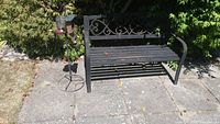 Metal black outdoor bench with scrollwork backrest and slatted seat shown in sunlight on concrete patio with surrounding greenery. Visible rust and weathering on bench.
