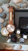 Image showing one wooden wall barometer with thermometer and several small clocks including round wall clock by Bulova and mantle clocks on stone hearth.