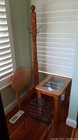 Wood coat rack with brass hooks, round finial top, folding wood table with circular top, black metal magazine rack with decorative pattern, and glass-topped wood end table grouped in corner of room.