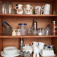 Three shelves filled with a variety of glassware, ceramic dinnerware, and kitchen accessories, showing wear and chips on some items.