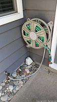 Wall-mounted beige plastic hose reel holding green garden hose attached to house exterior with visible rocks below.
