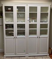 Front view of white IKEA Liatorp cupboard with four glass panel doors and solid lower panels, showing some contents inside.