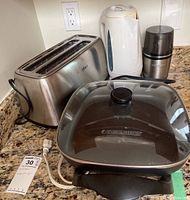 Black & Decker electric fry pan with glass lid, Oster stainless steel toaster, white electric kettle, Cuisinart coffee grinder on kitchen counter.