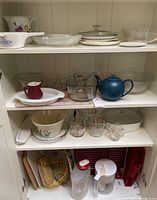 Front view of kitchen cabinet shelves showing multiple glass and ceramic items including bowls, measuring cups, teapot, and utensils stored inside cabinet.