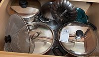 Photo showing various pots with glass lids, a steamer insert, and metal mesh strainer inside a drawer mostly assembled for sale.