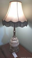 Full view of the table lamp illuminated, showing the tall cloth shade with fringe and the glass body on metal base.