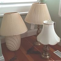 Three table lamps grouped showing varying bases and lampshades, placed on hardwood floor near window.