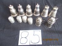 Top view of 12 assorted sterling silver salt and pepper shakers arranged in two rows on black cloth background showing different shapes and sizes.