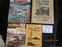 Four vintage historical books arranged on a black cloth, showing titles and cover illustrations: Yukon River Steamboats, Guide to Gold Panning in British Columbia, The Trail of 42, Lone Cove. Seller's lot number card '27' visible.