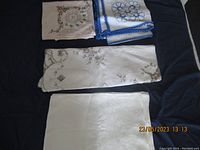 Four folded tablecloths displayed on dark surface: two smaller embroidered cloths on top, large beige embroidered cloth in middle, plain white cloth at bottom.