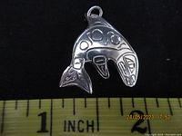 Front view of silver First Nations pendant showing carved design of stylized fish or orca with traditional motifs, placed next to ruler indicating size about 1.5 inches tall.