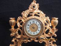 Front view of the Rococo style gold-tone clock showing the ornate scrollwork, face with numerals, missing hands, and two integrated candle holders.