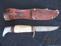 Photo showing full knife with natural handle, metal pommel and hollow ground blade next to embossed leather sheath featuring buffalo and Ruko branding.
