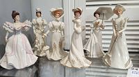 Group photo of six handcrafted Lenox lady figurines standing in a row on a shelf, showcasing their detailed period dresses with gold accents.