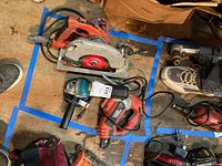 Milwaukee circular saw, Black and Decker mitre drill, Makita angle grinder, and Black and Decker corded tool arranged on floor inside marked blue tape boundary.