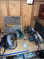 Four circular power saws placed on a wooden shelf in a shed. Two are gray/black, one is blue, all with power cords and some signs of use.