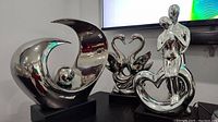 Front view of all three silver sculptures arranged on a table under a TV screen, showing abstract wave with sphere, swans forming a heart, and embracing couple figurine.