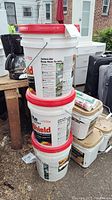Stack of three unopened white buckets with red lids of UltraShield Latex-ite driveway filler sealer with visible labels and nearby DAP sealant tubes and other items.