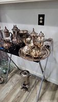 Collection of silverplate urns, pitchers and serving bowl set arranged on an oval serving tray displayed on a folding stand. Floor and wall outlet visible in background.