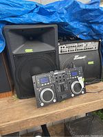 Photo showing large black speaker, amplifier, Gemsound speaker, and GenSound CMP-300 DJ controller laid out on wooden surface outdoors under a blue tarp.