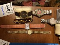 Six wristwatches arranged on wooden surface: Movado-style black-dial, USMC logo watch, Citizen silver watch, gold-tone mesh watch, Anne Klein pink strap, small gold-tone watch
