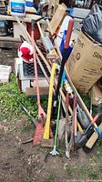 Photo showing a variety of yard tools including shovels, hoes, rakes, and brooms leaned against boxes and crates outdoors.