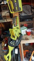 Photo of a yellow and black Ryobi cordless trimmer with handle, showing some dust and dirt marks, hanging in a workshop environment.